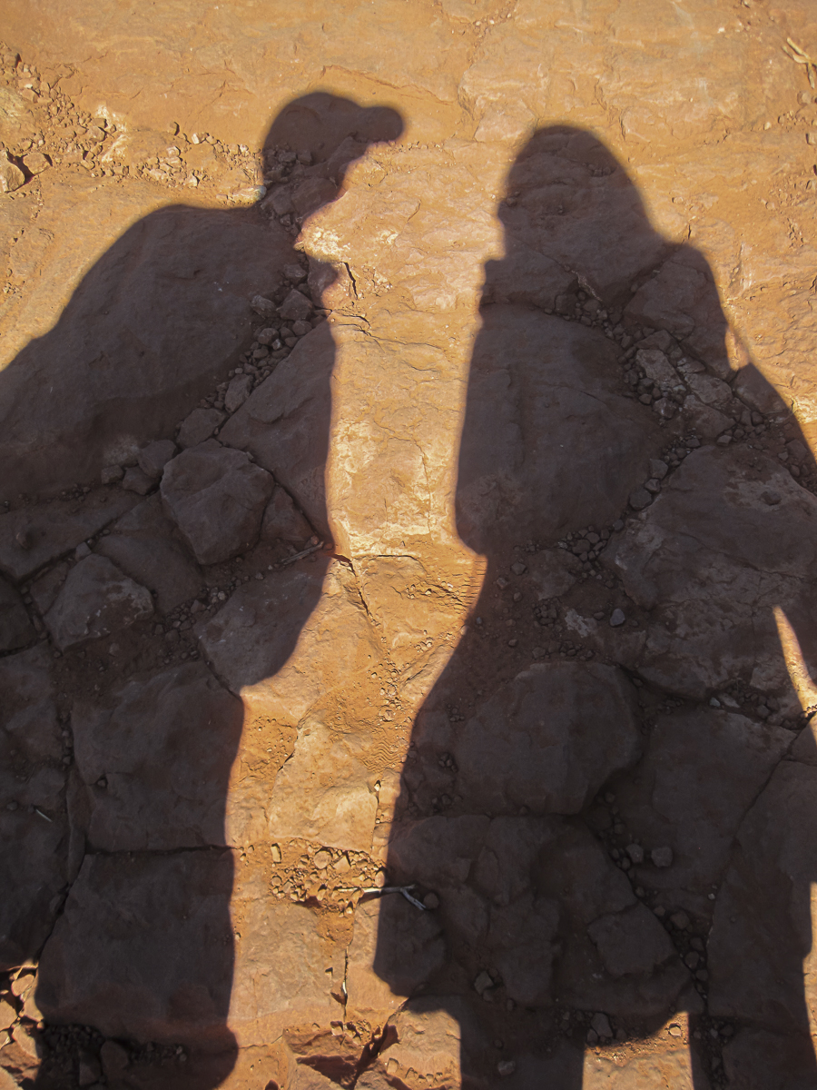 Shadows of three people cast on orange rocky ground in bright sunlight.