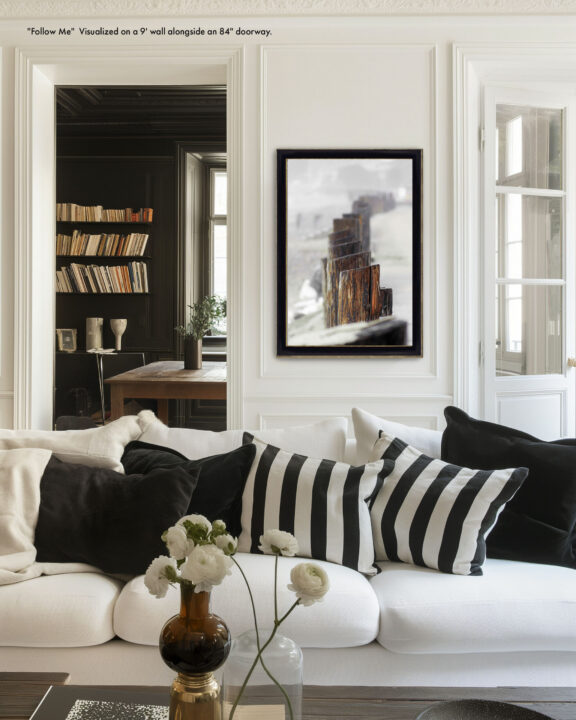 Bright living room with a white sofa and black-and-white striped pillows, glass vases and white flowers on the coffee table, doorway to a dark bookshelf-filled room in the background.